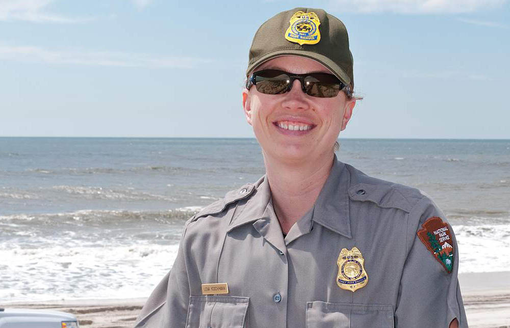 Lena Koschmann stands on a beach wearing NPS uniform and ball cap. She has a shield-shaped badge with an eagle perched on top pinned to her shirt.